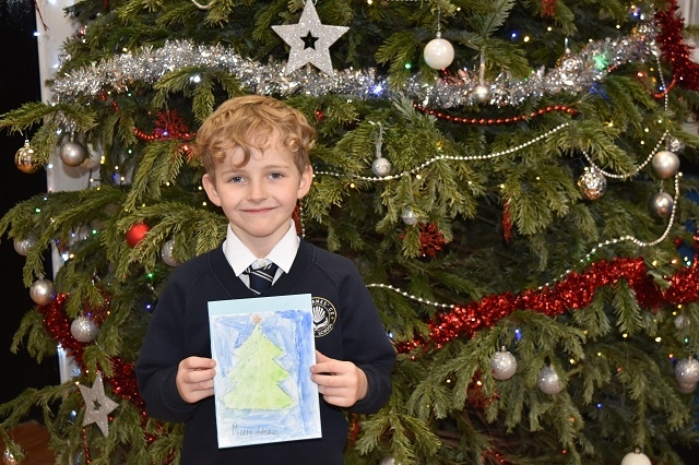 A boy from St James' CofE, Wardle, holds a Christmas card up in front of a decorated Christmas tree