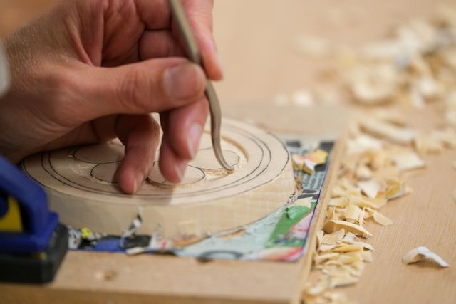  A close-up of someone's hand carving a flower design into a round block of wood using a pointed metal tool.