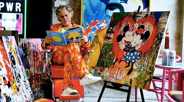 Johnny Zerox, dressed in a loud orange suit, reading a Disney book, surrounded by paintings, including a big artwork of Mickey and Minnie Mouse